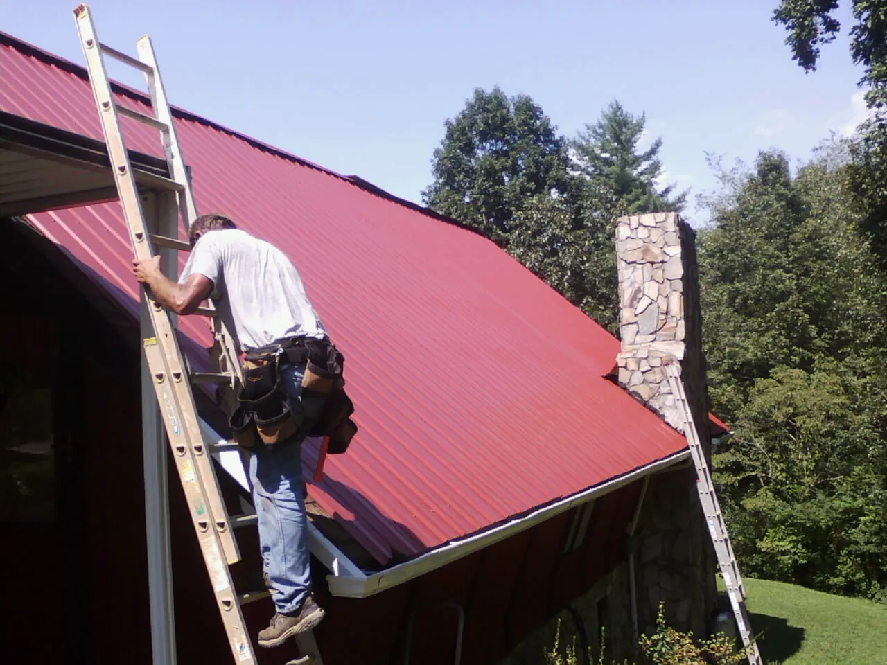 Roofer on a ladder beside a red metal roof and stone chimney — Soffit & Fascia Repair by Roofing Brothers in Fayette City